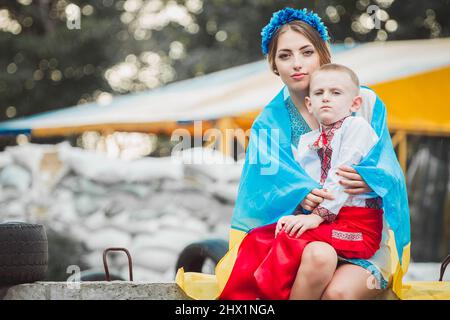 Die junge ukrainische Frau, die in Flagge gehüllt ist, sitzt mit dem Jungen in traditioneller Nationaltracht auf der Straßensperre vor dem Hintergrund von Sandsäcken. Konzept von russland Stockfoto