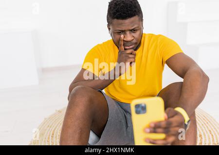 Schöner afroamerikanischer schwarzer Mann ernst zu gelb Smartphone mit kabellosen Kopfhörern. Selbstporträt. Aufgeregter junger afrikanischer Mann in Gelb Stockfoto