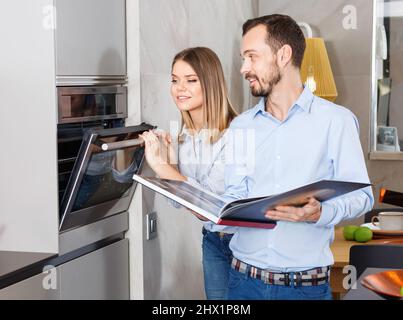 Junges, modernes Paar, das mit einem Rezeptbuch in einer stilvollen Küche in der Nähe des Backofens steht Stockfoto