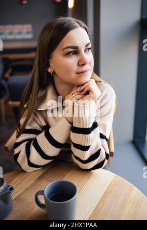 Ein braunäugiges Mädchen in einem Wollpullover lehnt sich an einen Tisch in einem Café und blickt aus dem Fenster. Eine junge Frau kam, um eine Tasse heißen duftenden Tee in einem zu trinken Stockfoto