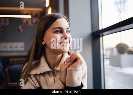 Ein braunäugiges Mädchen in einem Wollpullover lehnt sich an einen Tisch in einem Café und blickt aus dem Fenster. Junge Frau kam ins Café Stockfoto