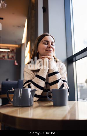 Ein braunäugiges Mädchen in einem Wollpullover lehnt sich an einen Tisch in einem Café und blickt aus dem Fenster. Eine junge Frau kam, um eine Tasse heißen duftenden Tee in einem zu trinken Stockfoto