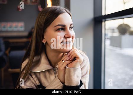 Ein braunäugiges Mädchen in einem Wollpullover lehnt sich an einen Tisch in einem Café und blickt aus dem Fenster. Junge Frau kam ins Café Stockfoto