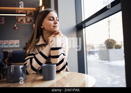 Ein braunäugiges Mädchen in einem Wollpullover lehnt sich an einen Tisch in einem Café und blickt aus dem Fenster. Eine junge Frau kam, um eine Tasse heißen duftenden Tee in einem zu trinken Stockfoto