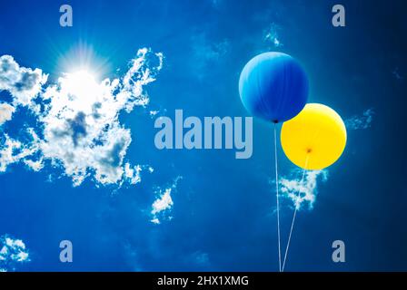 Zwei Luftballons in ukrainischen Flaggen-Farben über blauem Himmel und Sonne hinter den Wolken. Stockfoto