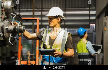 Maschinenbauingenieur, der in der Industrie und Fabrik mit reflektierender Schutzweste und weißem Helm arbeitet, Inspektor, der die industrielle Maschine im Stehen steuert und Stockfoto