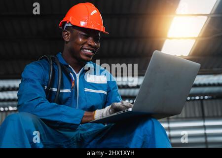 Glücklicher afrikanischer Elektroingenieur, der an einem Laptop arbeitet, indem er jährlich die elektrische Inspektion und Wartung im Werk überprüft und überprüft Stockfoto