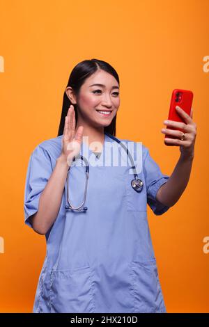 Lächelndes asiatisches Pflegepersonal in fernem Telemedizin-Videocall mit Patienten, während es eine Online-Konsultation auf orangefarbenem Hintergrund hat. Krankenschwester in der Klinik mit Stethoskop, winkt beim Videochat-Telefonkonferenz. Stockfoto