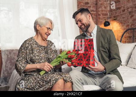 Die glückliche Oma erhält von ihrem bärtigen Enkel mittleren Alters eine Geschenktüte und einen Strauß. Hochwertige Fotos Stockfoto