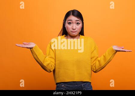 Verwirrt ungewiss asiatische Frau zuckte unsicher, während das Tragen gelben Pullover auf orangefarbenem Hintergrund. Ahnungslos unsicher junge Erwachsene Mädchen spreizen Hände seitwärts, während Gefühl Zweifel. Stockfoto