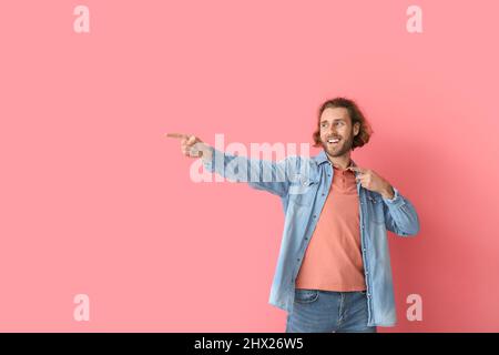Hübscher junger Mann in stilvoller Kleidung, der auf etwas auf rosa Hintergrund zeigt Stockfoto