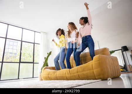 Drei junge Erwachsene Freundinnen, die Spaß haben, gemeinsam auf dem Sofa zu Hause zu tanzen Stockfoto