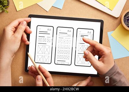 Zwei UI-/UX-Front-End-Designer arbeiten zusammen und nutzen ein Tablet-Gerät, um mobile Anwendungswireframes auf dem Schreibtisch zu analysieren. Stockfoto