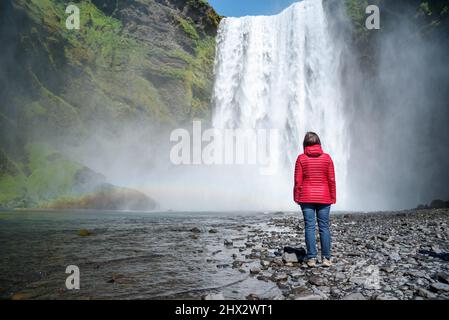 Frau, die an einem sonnigen Sommertag am Fuße eines mächtigen Wasserfalls steht Stockfoto