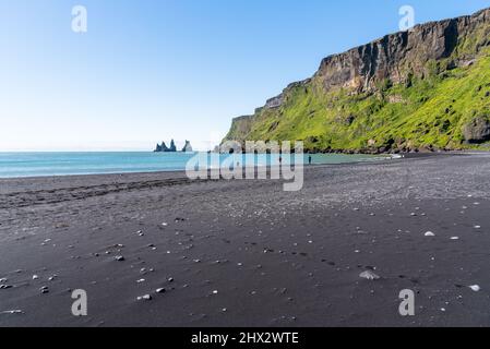 Menschen, die sich am Ufer eines schwarzen Sandstrands in Island Vergnügen. Fußabdrücke im Sand sind im Vordergrund sichtbar. Stockfoto