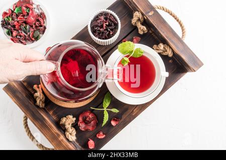 Tee Ceremonia . eine Frauenhand gießt duftenden roten Hibiskustee in eine Tasse Minzblätter. Draufsicht auf das Holztablett mit Morgentee Stockfoto