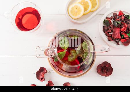 Draufsicht auf die Teekannen mit frisch gebrühtem Hibiskusblütentee und Minzblättern. Zitrone zur Verbesserung von Geschmack und Vitaminen. Weißer Holztisch Stockfoto