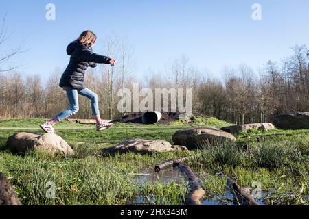 Ein junges Mädchen springt zwischen den Trittsteinen auf einem Hindernisparcours in einem Park, bei strahlender Frühlingssonne, in einem Wald in Diemen, Niederlande. Stockfoto