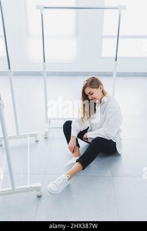 Verspielte selbstbewusste Frau in einem stilvollen weißen Baumwollhemd, schwarzer Jeans, weißen Leder-Sneakers, lässigem Stil. Blonde Frau auf einem grauen Rückenverlaufe Stockfoto