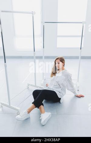 Verspielte selbstbewusste Frau in einem stilvollen weißen Baumwollhemd, schwarzer Jeans, weißen Leder-Sneakers, lässigem Stil. Blonde Frau auf einem grauen Rückenverlaufe Stockfoto