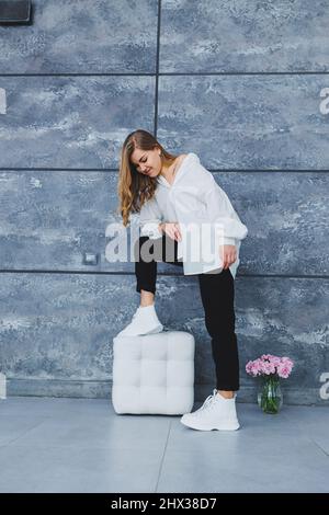 Verspielte selbstbewusste Frau in einem stilvollen weißen Baumwollhemd, schwarzer Jeans, weißen Leder-Sneakers, lässigem Stil. Blonde Frau auf einem grauen Rückenverlaufe Stockfoto