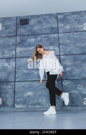 Verspielte selbstbewusste Frau in einem stilvollen weißen Baumwollhemd, schwarzer Jeans, weißen Leder-Sneakers, lässigem Stil. Blonde Frau auf einem grauen Rückenverlaufe Stockfoto