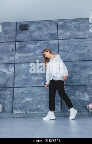 Verspielte selbstbewusste Frau in einem stilvollen weißen Baumwollhemd, schwarzer Jeans, weißen Leder-Sneakers, lässigem Stil. Blonde Frau auf einem grauen Rückenverlaufe Stockfoto