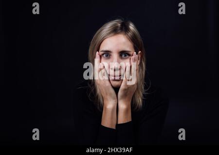 Ängstliche junge Frau, die mit Händen auf den Wangen in einem schwarzen Kleid auf schwarzem Hintergrund auf die Kamera schaut. Opfer von körperlicher und psychischer Misshandlung Stockfoto