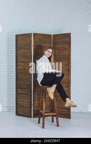 Eine fröhliche, schöne junge Frau mit einem Lächeln in einem modischen Outfit mit weißem Hemd, beigefarbenen Lederschuhen und schwarzer Hose sitzt auf einem Holzstuhl. Stockfoto