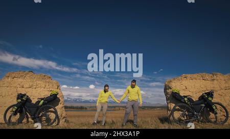 Der Mann und die Frau reisen mit Bikepacking auf gemischtem Terrain. Die beiden Personen reisen mit Fahrradtaschen. Sportbekleidung in grün-schwarzen Farben. Schneebedeckter Bergbogen. Stockfoto