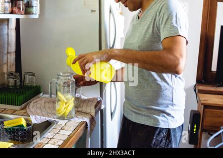 Junger Mann, der zu Hause in der Küche steht und Wasser in einen Glaskrug mit Zitronenscheiben gießt, Lifestyle-Konzept. Stockfoto