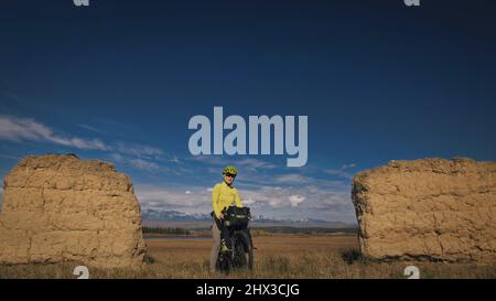 Die Frau reist auf gemischtem Terrain mit Bikepacking. Die Reise des Reisenden mit Fahrradtaschen. Sport Bikepacking, Fahrrad, Sportbekleidung in grün-schwarzen Farben. Schneebedeckter Bergbogen. Stockfoto