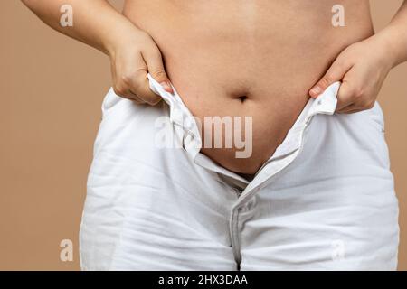 Nahaufnahme einer Dame mit dickem plumpigen Bauch, die versucht, weiße Hemden auf beigem Hintergrund zu verreißen. Viszerales Fett. Körper positiv. Plötzliche Gewichtszunahme. Fest Stockfoto