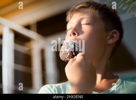Junge Teenager isst Schokoladenpopsicle. Geschlossene Augen vor Freude. Eis in der Hitze. Stockfoto