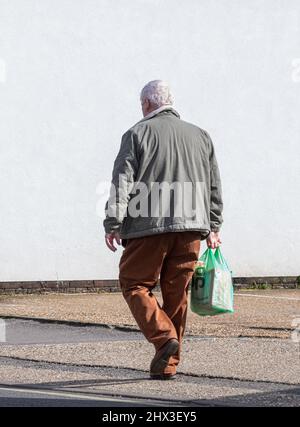 Älterer Mann, der aus dem Geschäft oder Geschäft zurückkommt und eine Co-op-Supermarkttasche aus Kunststoff trägt, Großbritannien Stockfoto