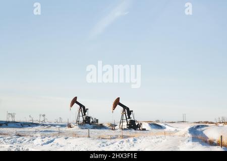 Zwei Erdölpumpen-Wagenheber hinter einem Zaun in einer landwirtschaftlichen Winterlandschaft auf den Prärien Stockfoto