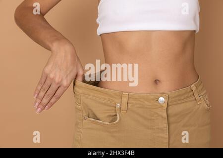 Nahaufnahme von Frau Körper in großen Hosen mit der Hand ziehen sie schlanke Form auf körperfarbenen Hintergrund zu zeigen. Gewichtsverlust durch die Aufrechterhaltung der Ernährung und Bewegung Stockfoto