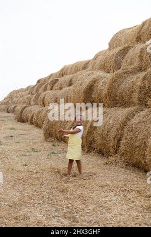 Porträt eines niedlichen kleinen Mädchens, das neben einem Haufen gerollter Heuhaufen auf dem Heufeld steht und die Kamera mit der Hand in der Mundspüllung auf etwas zeigt. Haben Stockfoto