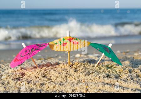 Drei kleine Papier-Cocktail-Schirme stehen im Sand am Sandstrand aus nächster Nähe. Stockfoto