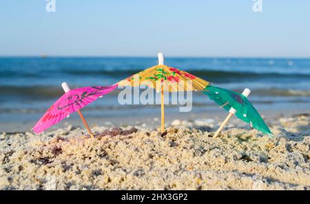 Drei kleine Papier-Cocktail-Schirme stehen im Sand am Sandstrand aus nächster Nähe Stockfoto