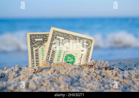 Zwei ein-Dollar-Scheine, die zur Hälfte im Sand an der sandigen Küste aus nächster Nähe vergraben sind. Stockfoto