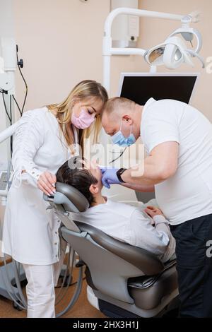 Der Zahnarztarzt schaut auf die Zähne des Patienten und hält zahnärztliche Instrumente in Mundnähe. Der Assistent hilft dem Arzt. Sie tragen weiße Uniformen Stockfoto