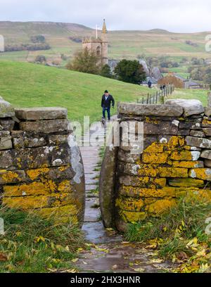 Ein Mann, der seinen kleinen Hund auf einem Fußweg in der Bealer Bank in Hawes, Wensleydale, zu einem Squeeze Stile führt Stockfoto