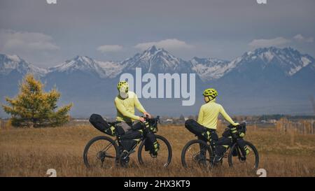 Der Mann und die Frau reisen mit Bikepacking auf gemischtem Terrain. Die beiden Personen reisen mit Fahrradtaschen. Sport Bikepacking, Fahrrad, Sportbekleidung in grün-schwarzen Farben. Schneebedeckte Berge. Stockfoto