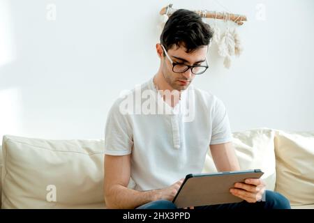 Porträt eines freiberuflichen Mannes, der zu Hause immer in Kontakt ist. Hübscher, fokussierter junger Mann, der sein Smartphone benutzt, während er auf der Couch sitzt. Stockfoto