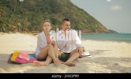 Das fröhliche Liebespaar hält und isst Wassermelonenscheiben am tropischen Sandstrand. Romantische Liebhaber zwei kaukasische Menschen verbringen das Sommerwochenende im Freien. Mütze, Rucksack weißes Hemd Strandmode. Stockfoto