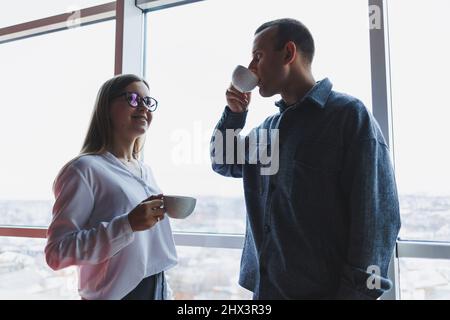 Gutaussehender männlicher Geschäftsmann und junge Geschäftsfrau stehen und reden am Morgen vor der Arbeit im Bürogebäude, sie trinken Stron Stockfoto