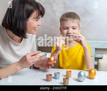 Mama und ein kleiner blonder Junge malen Eier mit Bürsten für die Osterferien in der heimischen Küche. Osterurlaub mit der Familie. DIY Ostereier Konzept. Stockfoto