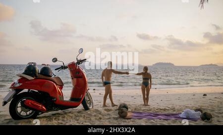 Motorroller Road Trip. Schönes Paar auf rotem Motorrad in weißen Kleidern am Sandstrand. Hochzeit gerade verheiratete Menschen, die in der Nähe von tropischen Palmen, Meer. Flitterwochen am Meer. Asien Thailand. Motorradmiete. Stockfoto