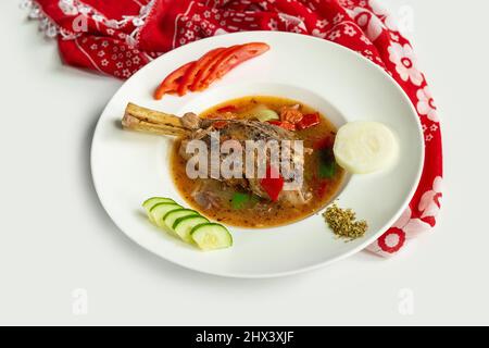 Geschmorte Lamb Shanks mit Sauce und Tomaten, Gurken, Rettich in einem Gericht isoliert auf bunten Tischtuch Draufsicht auf grauem Hintergrund Stockfoto